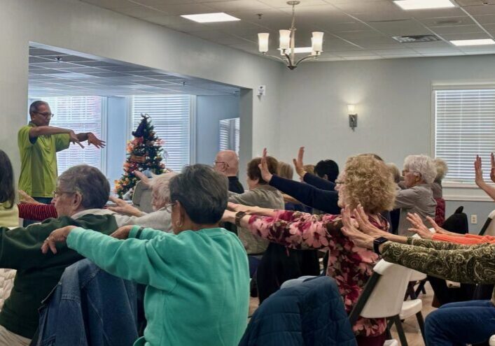 Group of elderly people exercising with arm stretches indoors.