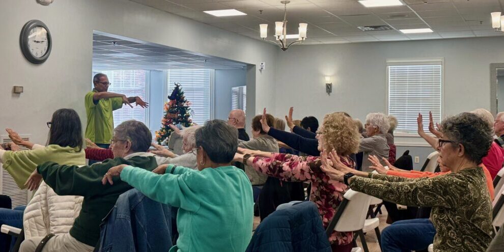 Group of elderly people exercising with arm stretches indoors.